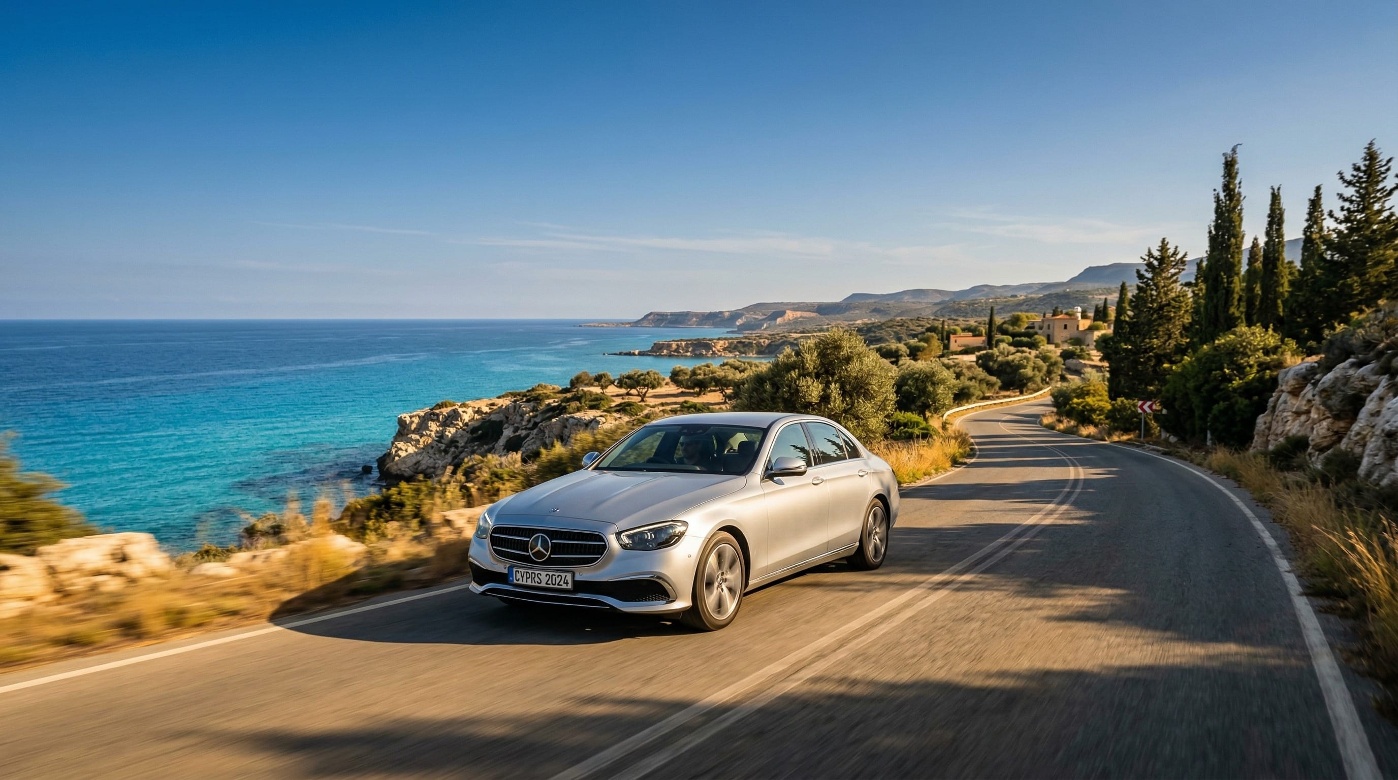 Ένα σύγχρονο ασημί αυτοκίνητο οδηγεί σε γραφικό παραλιακό δρόμο στην Κύπρο, με τιρκουάζ Μεσόγειο θάλασσα, ασβεστολιθικούς βράχους και ελαιόδεντρα στο βάθος κάτω από ζεστό χρυσαφί απογευματινό φως.