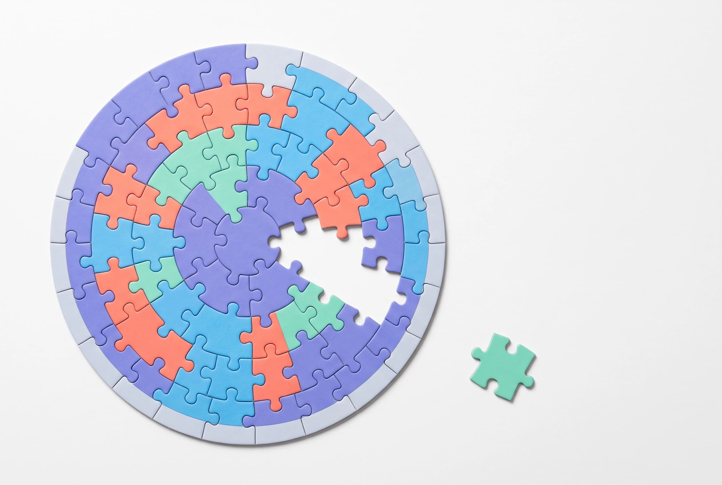 An overhead view of a nearly complete circular puzzle with one mint-green piece missing and sitting apart from it.