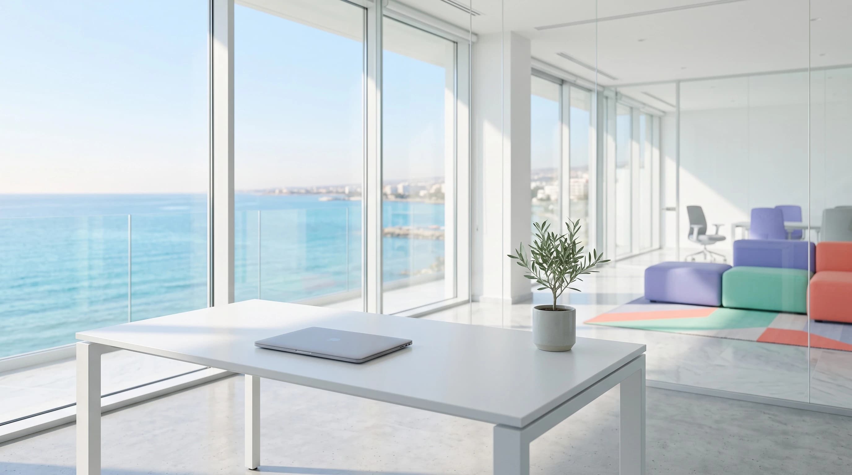Bright modern Limassol office with floor-to-ceiling windows overlooking the Mediterranean, a white desk with a closed laptop and a small olive plant.