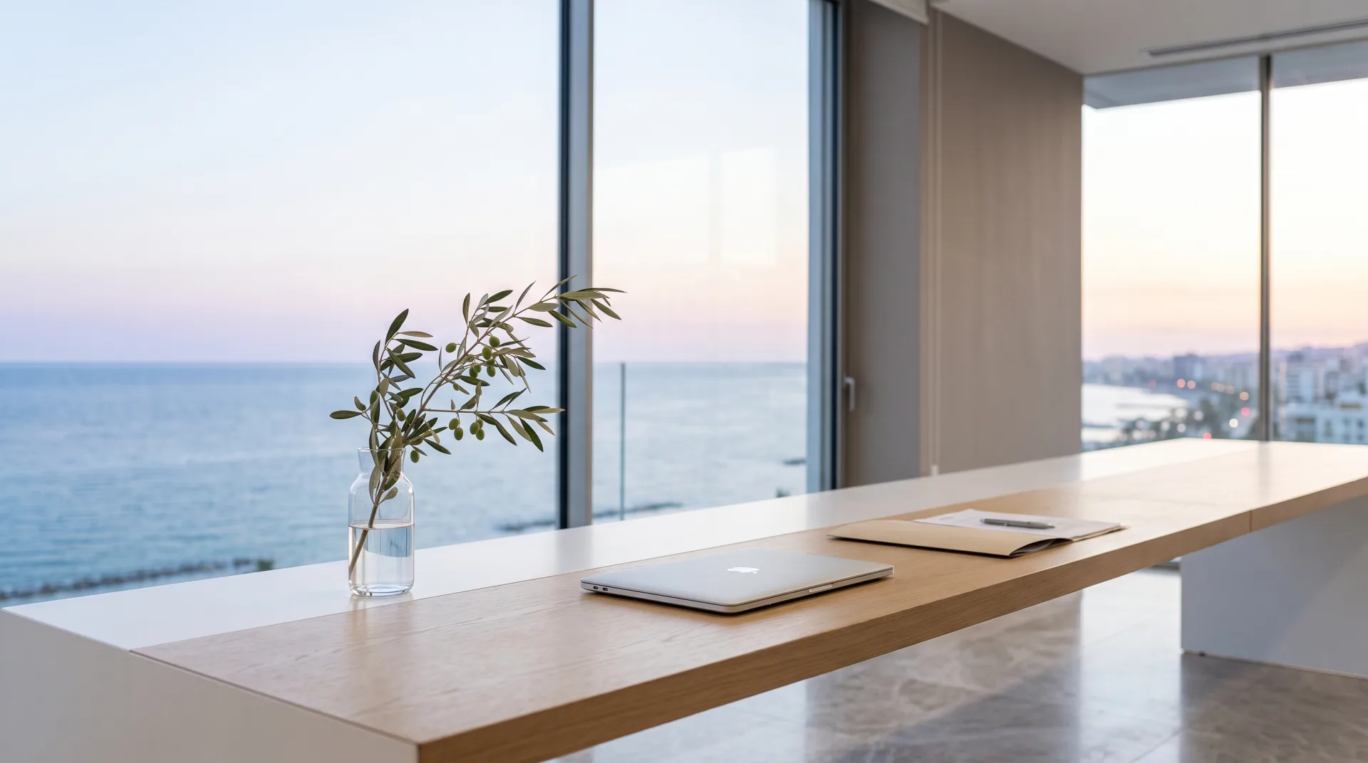 Quiet Limassol fund office at first light, a long white desk with a closed laptop, a single olive branch in a glass and the sea beyond the windows.