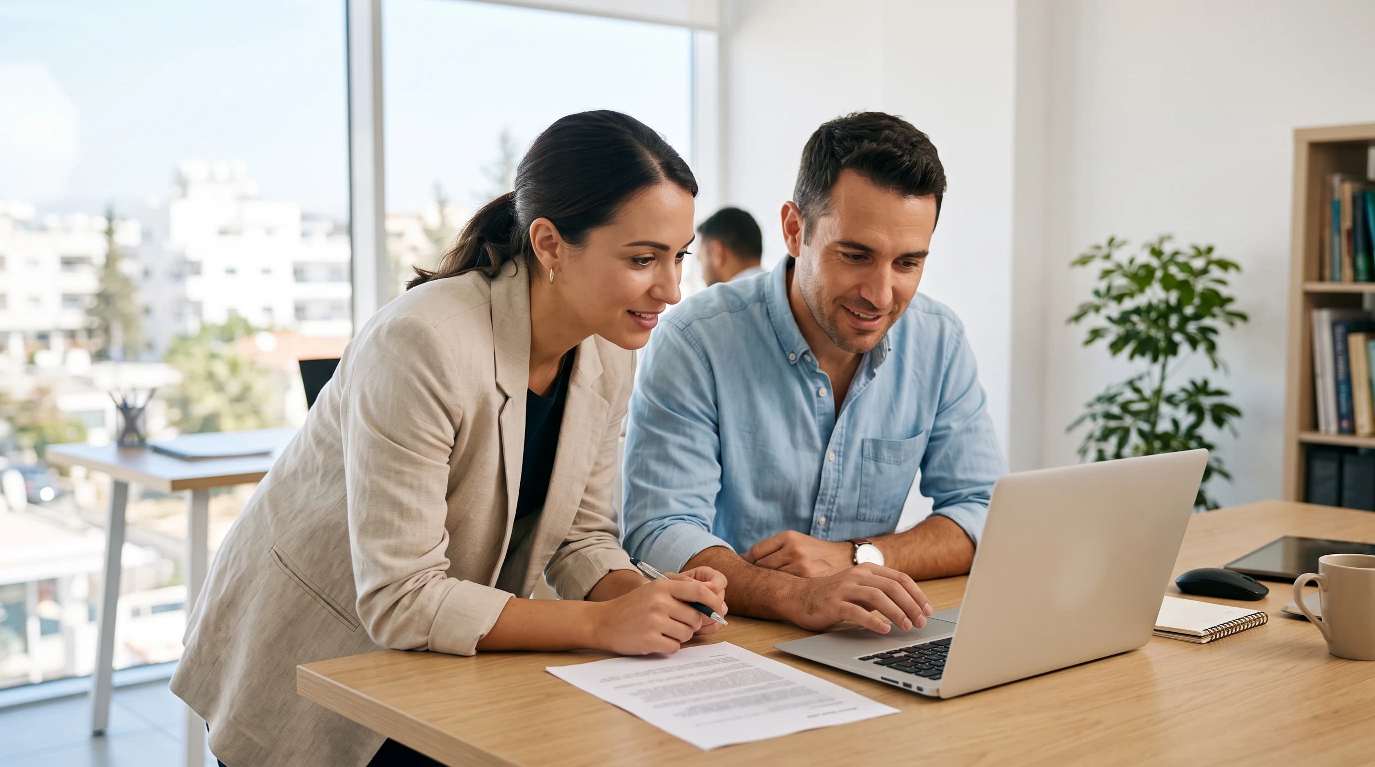 Two developers at a white desk in a bright Limassol office, one laptop open and a printed contract beside it.